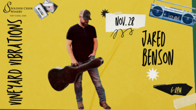 Jared Benson stands with a guitar case in hand, wearing a ballcap, dark shirt, jeans, and boots. The background is yellow with black and white cutout elements, including tape strips, stars, and a boombox. Text reads: “Vineyard Vibrations,” “Nov. 28,” “Jared Benson,” and “6–8PM.” Soldier Creek Winery logo in top left.