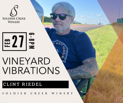 Promotional graphic for Vineyard Vibrations at Soldier Creek Winery featuring Clint Riedel on February 27 from 6–8PM. Clint is seated in a lawn chair near a highway, wearing sunglasses and a blue t-shirt with tattoos visible on his arm. Blue sky and road signs are in the background.