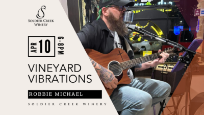 Vineyard Vibrations live music event at Soldier Creek Winery featuring Robbie Michael on April 10, 6–8pm. Photo shows a bearded man playing an acoustic guitar and singing into a vintage-style microphone.