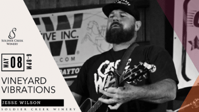 Vineyard Vibrations live music event at Soldier Creek Winery featuring Jesse Wilson on May 8, 6–8pm. Black-and-white photo shows a bearded man in a cap playing guitar and singing outdoors.